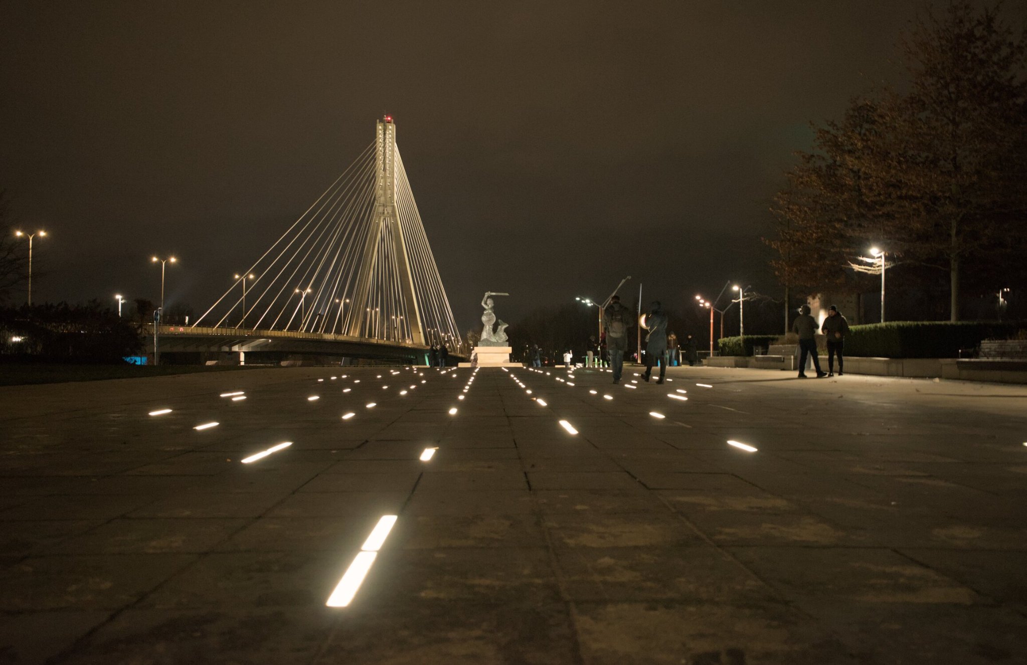 a walkway illuminated with stripes of light in the ground, and a bridge in the distance... the warsaw... mascot(?) is situated towards the central point of the frame, folks scattered around the place