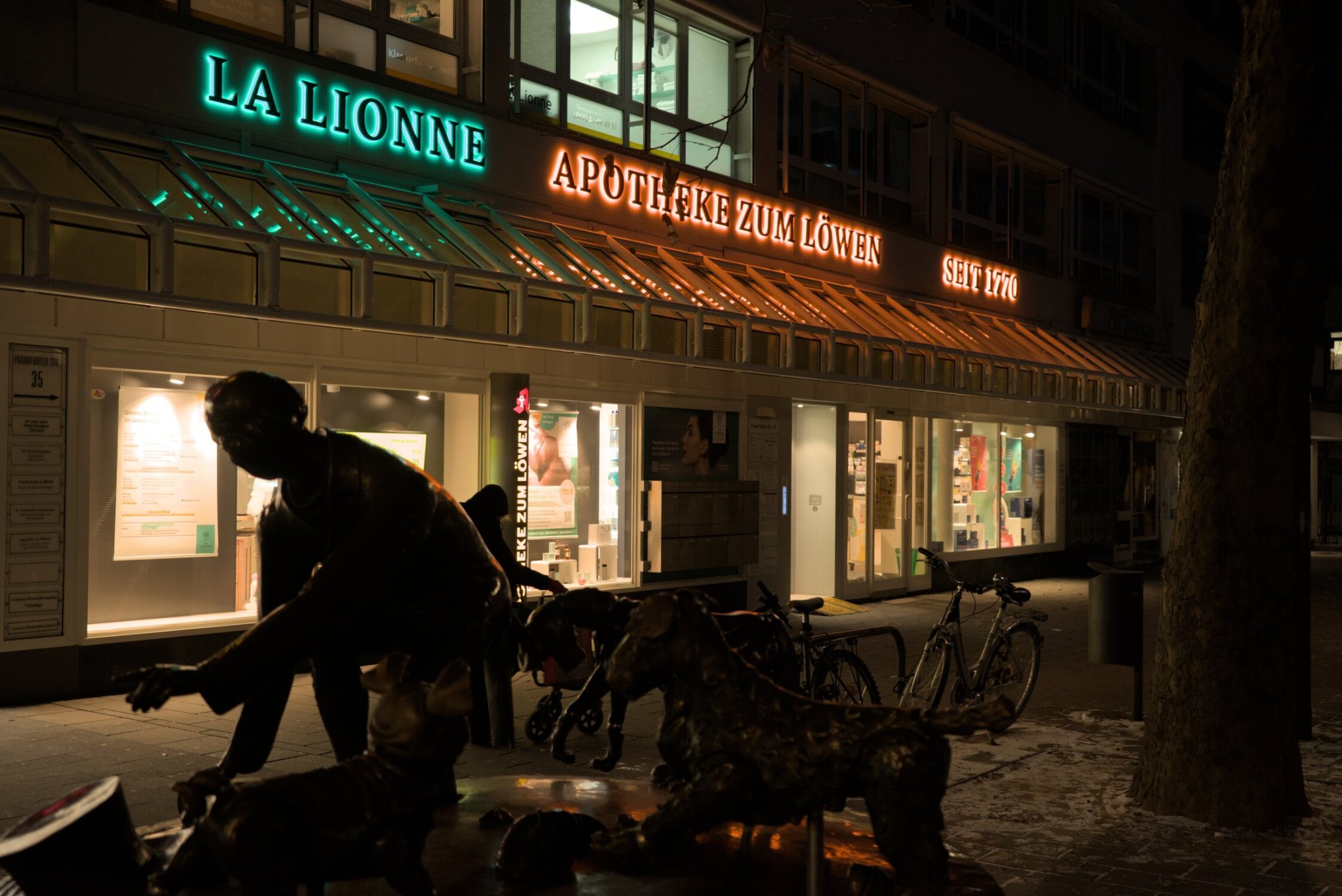 a few shops with red and green signs lie in the background, with a person pushing a cart obscured by a bronze statue of a person and dogs in the foreground