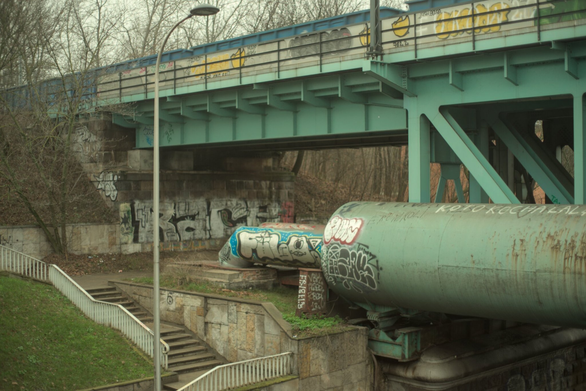a grafittied train bridge, with one currently passing overhead, stairs and grass connect it to the road below