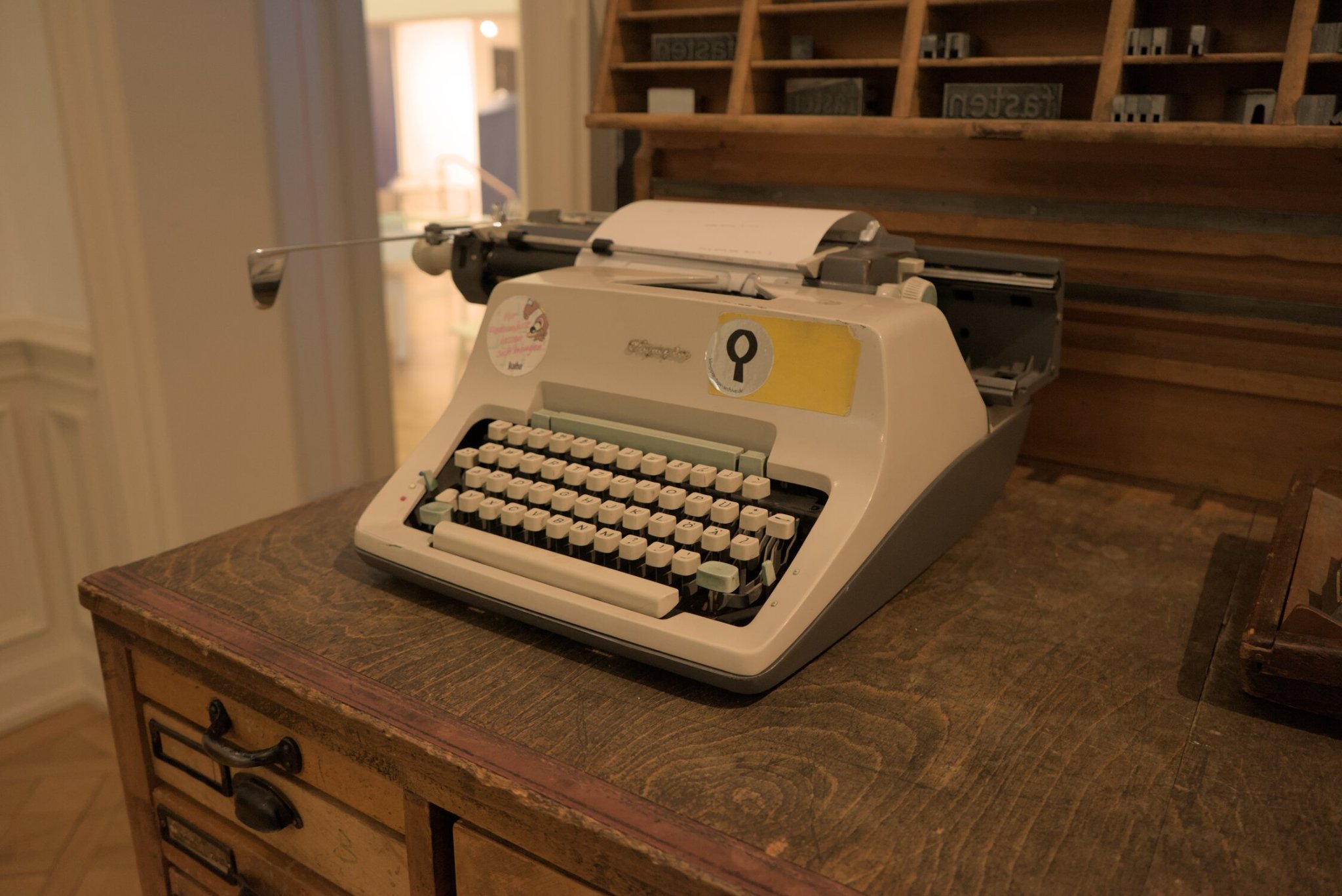 a typewriter with QWERTZ layout, the shift keys being teal, and the others being a creamy white, a few stickers adorn its face and a sheet of paper is loaded with some illegable text printed upon it