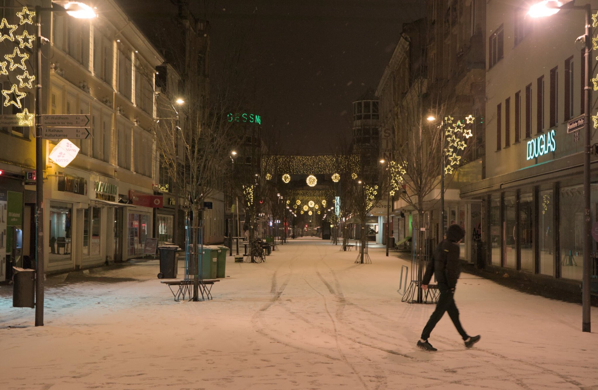 a market street, winter celebrations concluded, decorations still adorning... a person walks hurridly past as it gently snows