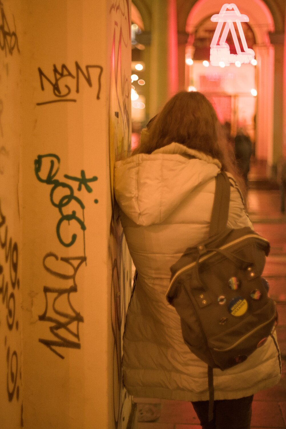 @xyla@shitpost.trade leaning agains a graffitied wall, taking a picture of the scene beyond, bundled in a winter coat, waiting for just the right moment... the scene is very warm, having the double whammy of sodium vapour lighting and my thorium super takumar lens
