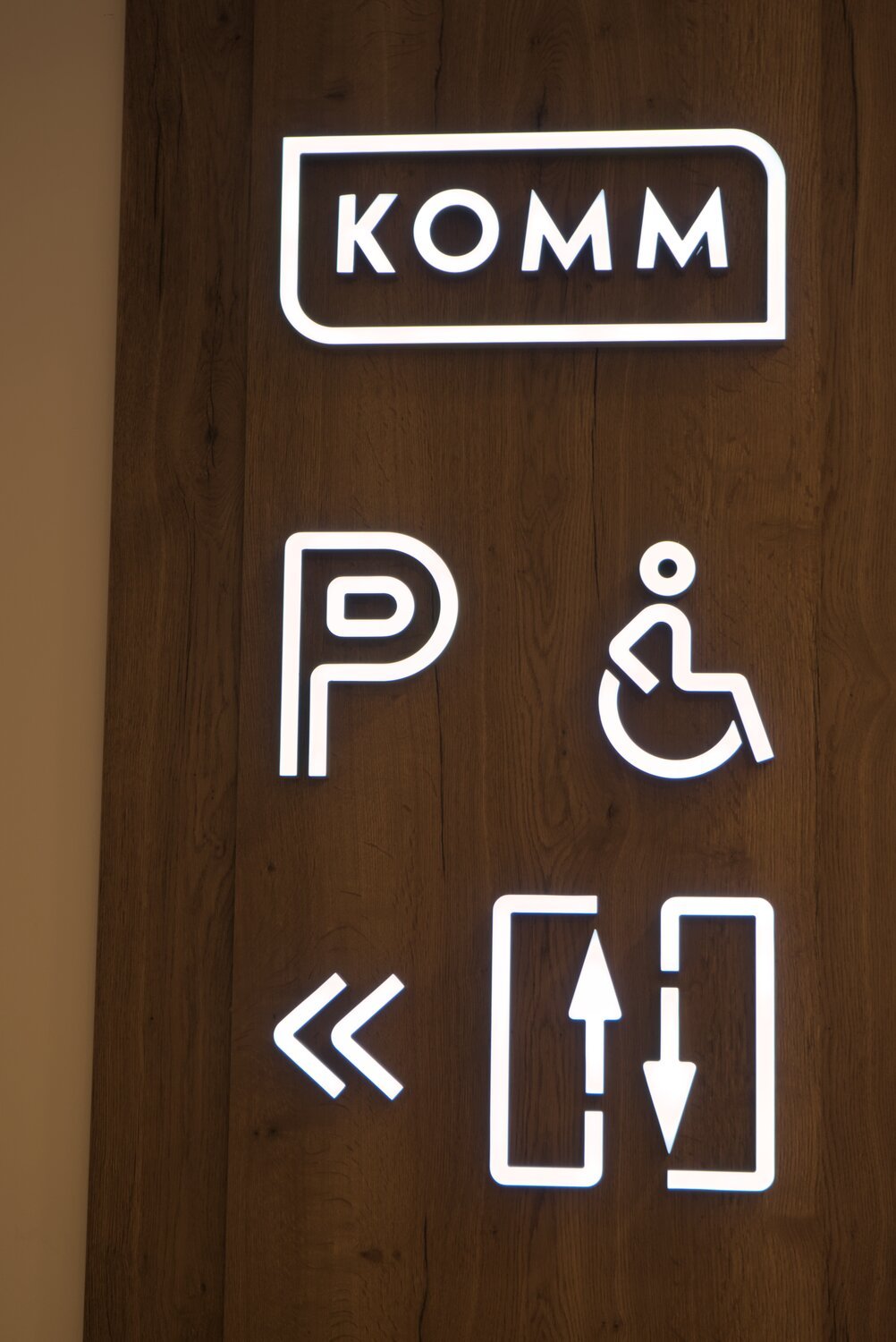 a navagation board of sorts for 'KOMM', with white illuminated lettering on a woodgrain panel, showing directions for parking, accessability, and escalators 