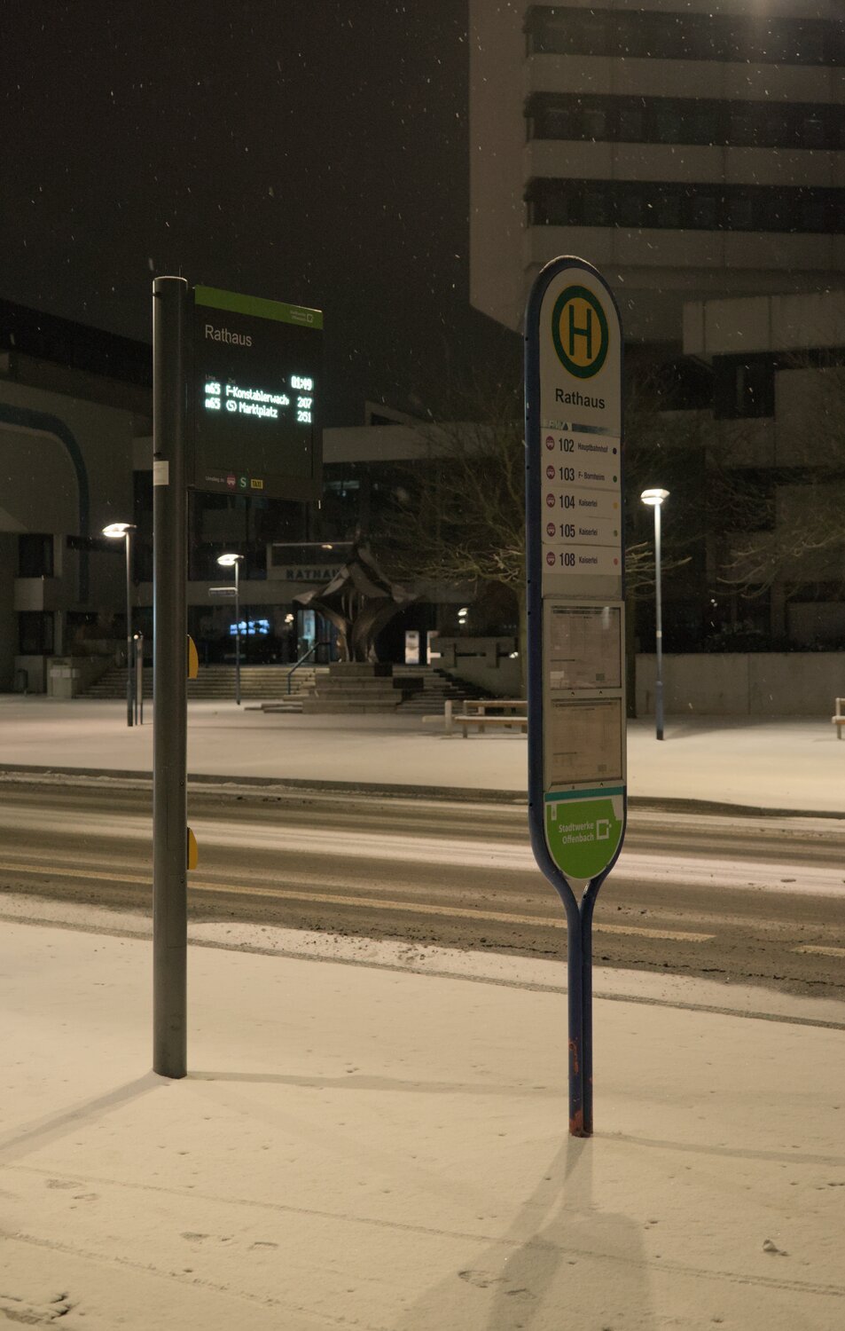 a bus stop and departures board for the rathaus, it's snowing gently, and the rathaus in the background has turned off its lighting for the night