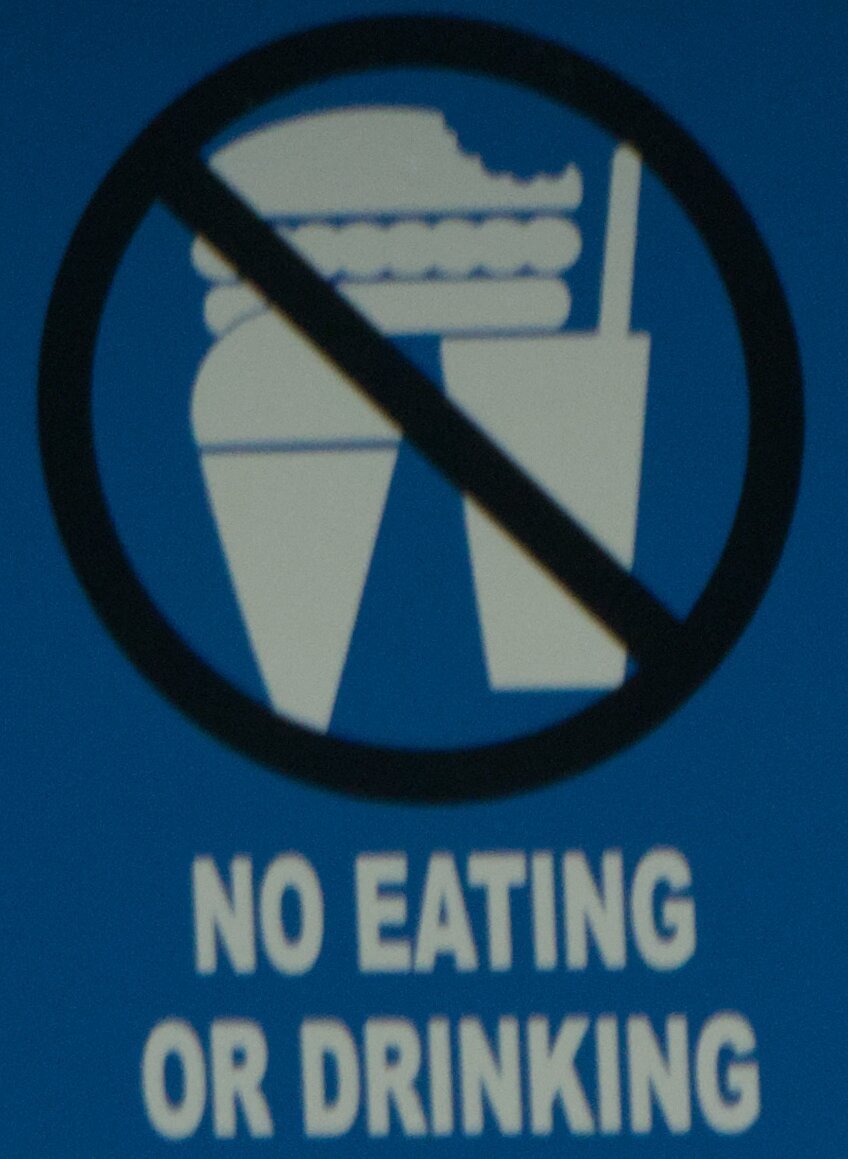 a sign with white silhouettes of a sandwich, ice cream cone and drink, all crossed through with a 'prohibited' symbol, the sandwich has a bite akwardly eaten from the top bun
the text on the sign reads 'no eating or drinking'