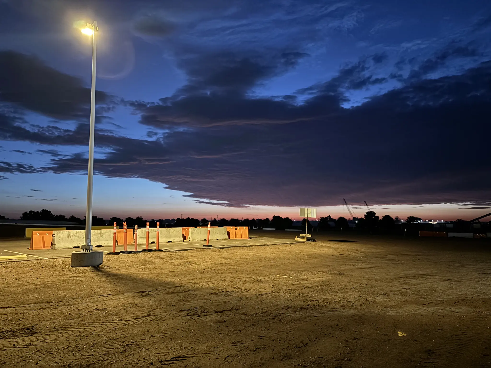 a sunrise as viewed from a dirt lot, a lightpole illuminates barricades on the pavement beyond