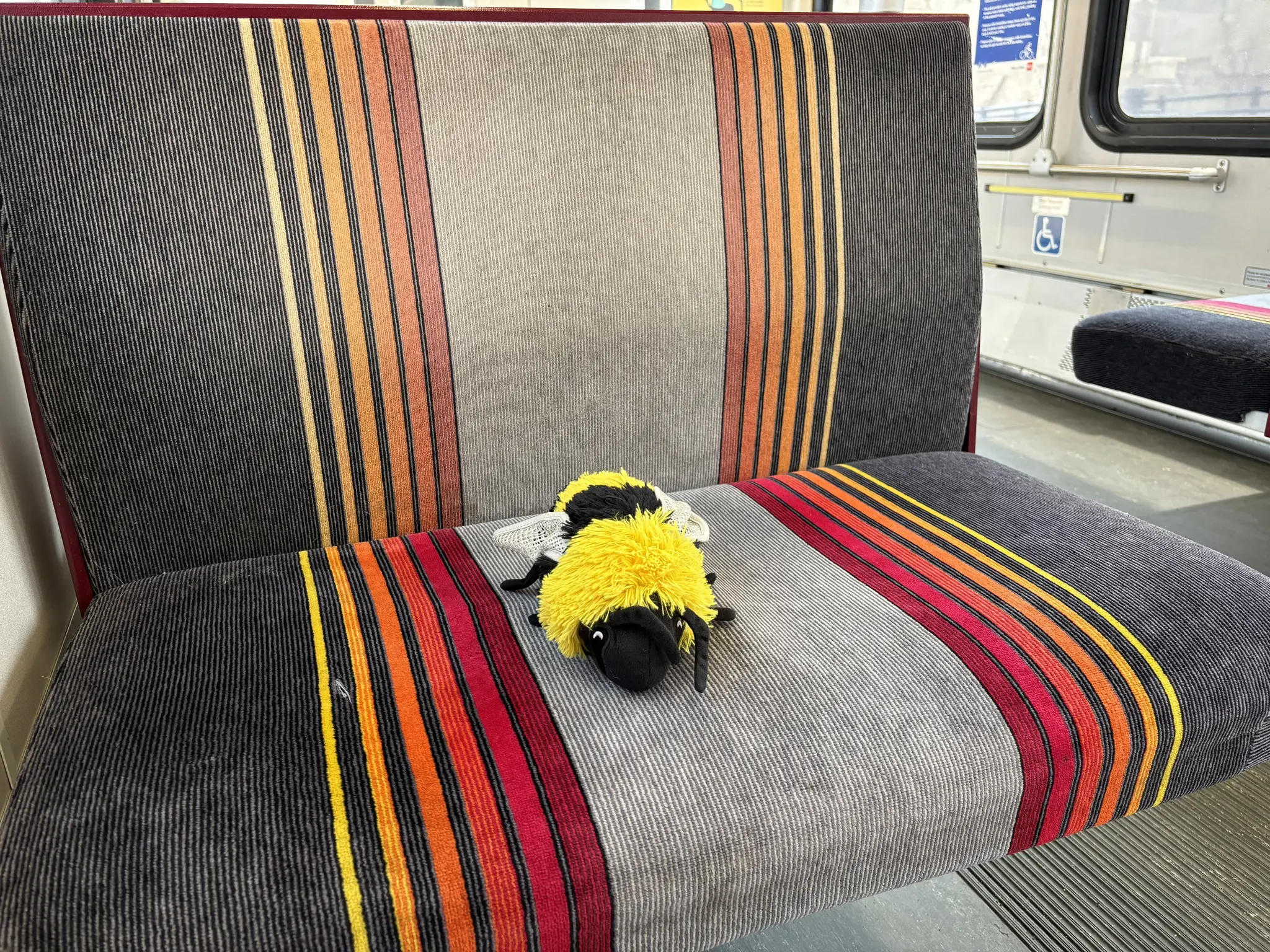 here, we have a plush bee, resting on a bench seat far too large for it!
the seat has a large dark grey stripe, then alternating yellow fading to red and drak gray, with a wide light gray stripe in the middle