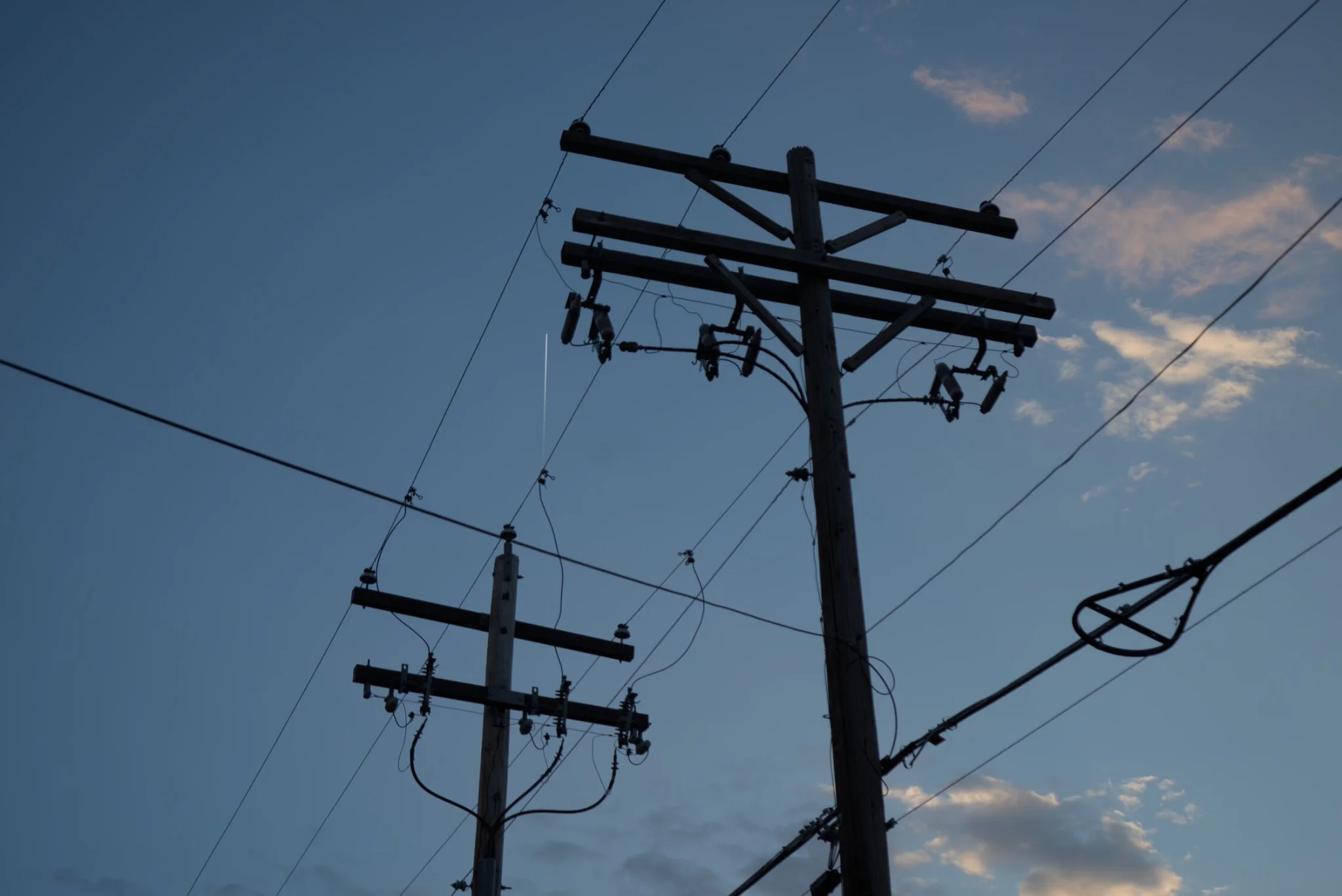 silhouettes of two power poles in front of a partly cloudy sunset sky, there is a plane contrail visible roughly center frame