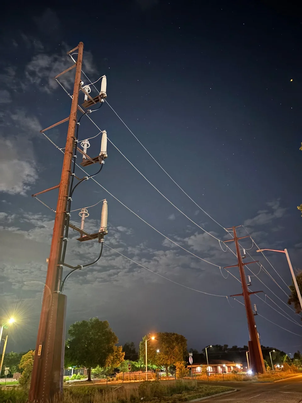 two big power poles, the view looking up at them towards the night sky, some clouds can be seen to the left, with stars to the right and trees and light poles to the bottom