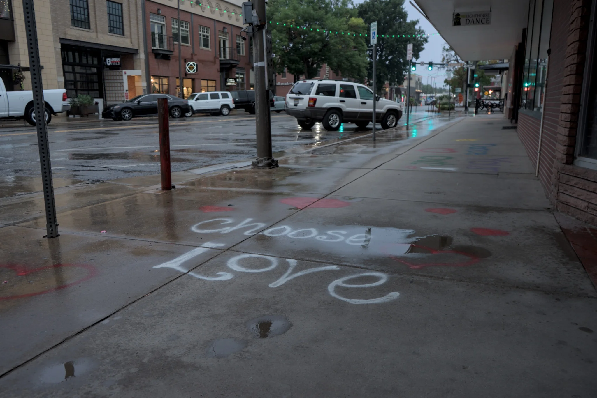 sidewalk art that says 'Choose Love' in white with red hearts surrounding
much of the image is slightly blurry and there is a puddle over the 'se' in choose
it's quite rainy in the scene