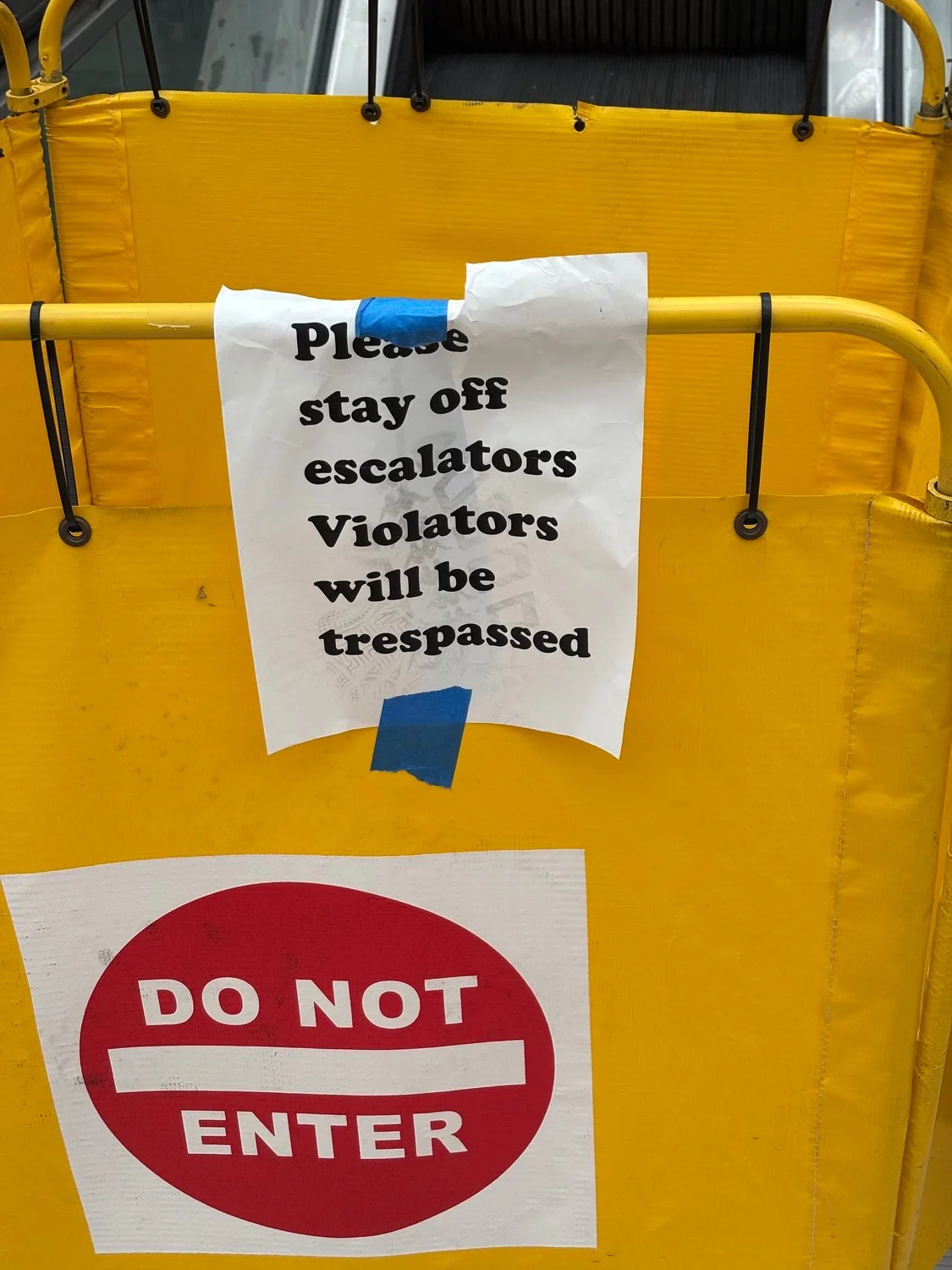 A yellow barricade with two signs attached
Upper sign reads: "Please stay off escalators Violators will be trespassed"
Lower sign reads: "Do Not Enter"