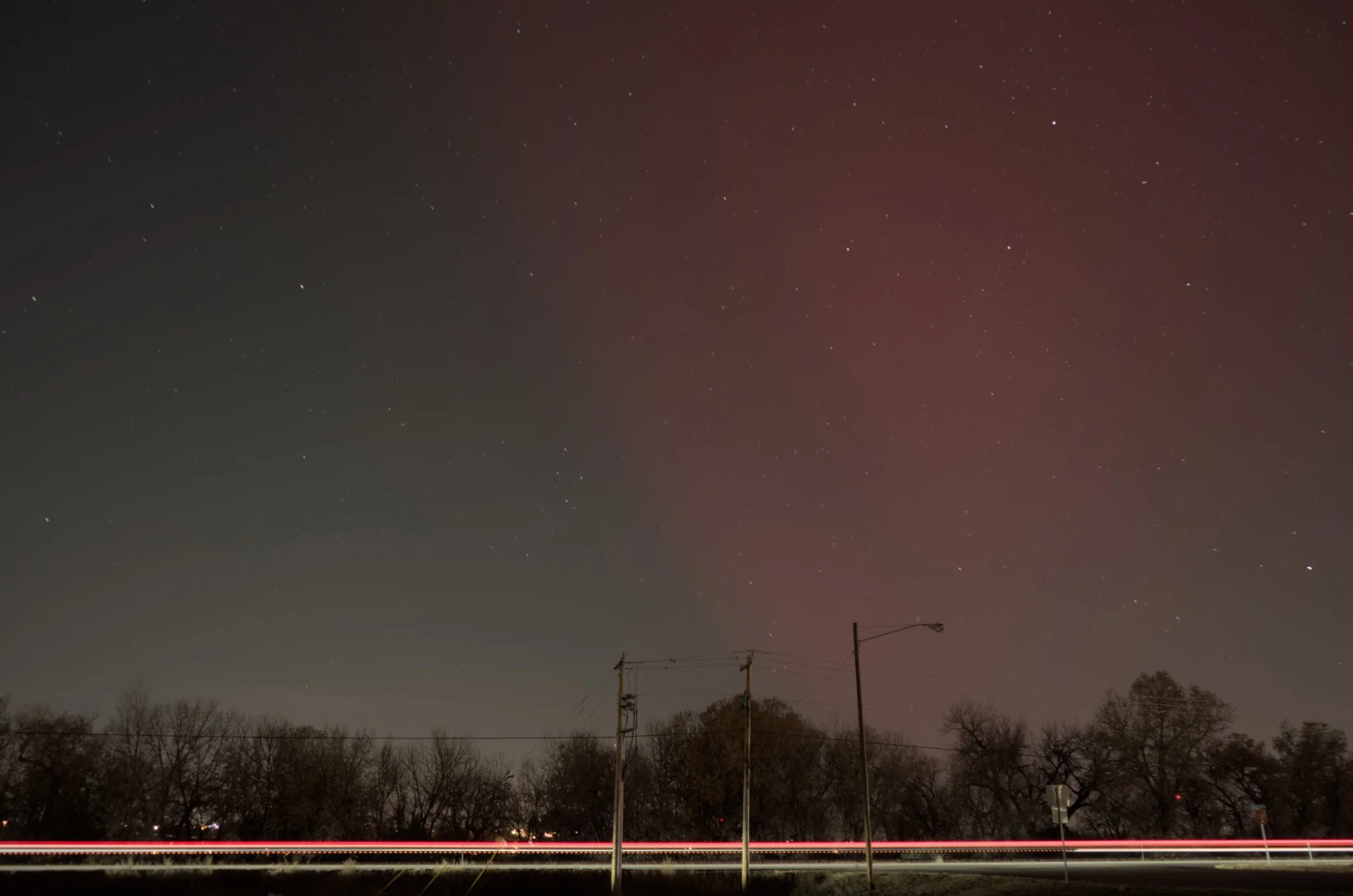 a massive streak of red aurora visible in the central, light polluted sky, with stars faintly visible
streaks of a car's head and taillights are visible at the bottom of the frame