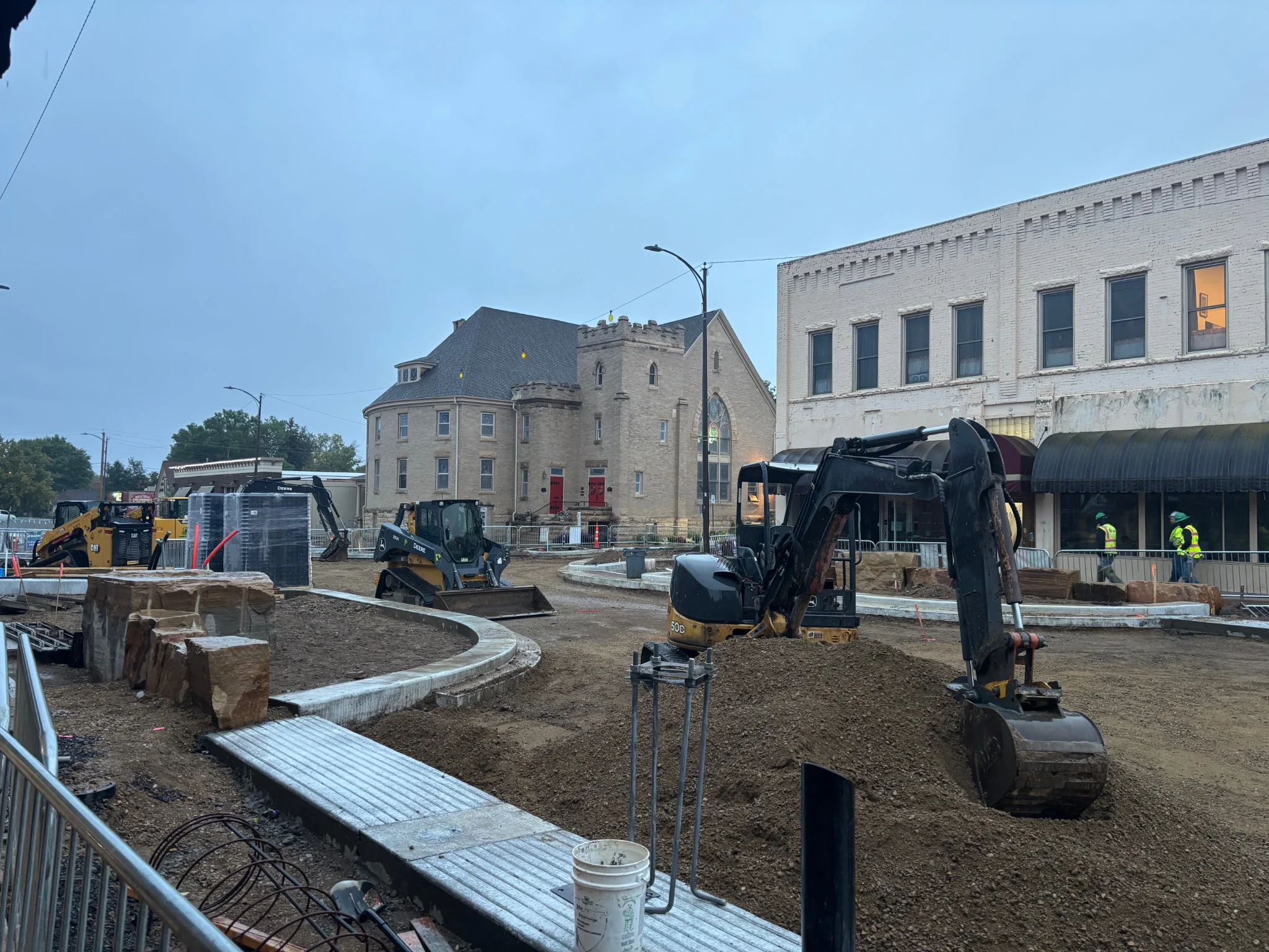 a road under renovation, digging trucks are in the scene, with a church of some sort in the background
there are construction workers walking away from the viewer on the right hand edge of the frame