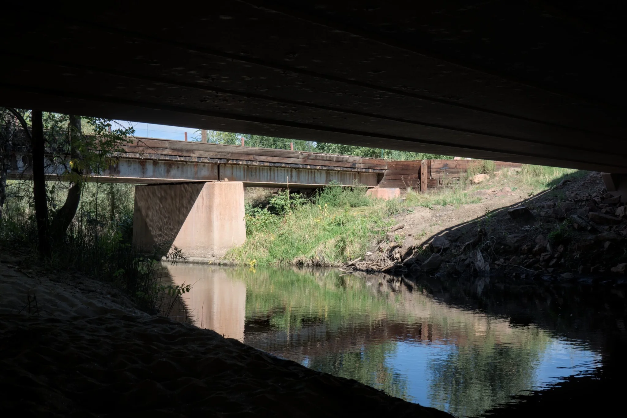 a river, with some train tracks behind it
the viewer is under a bridge and in shadow, with the bridge in sunlight