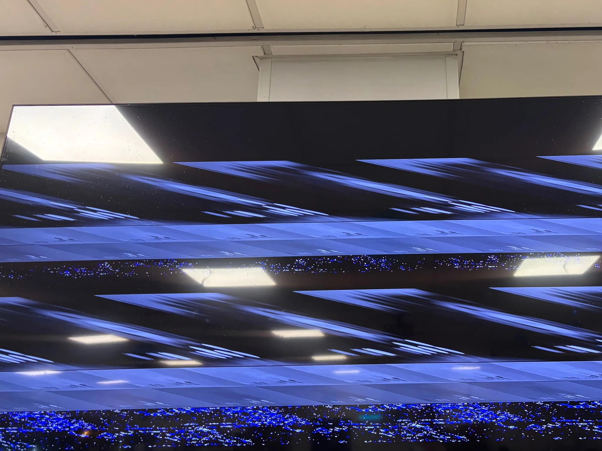 a glossy black television, upon its display is something that is supposed to mean something, but is indecipherable due to corruption
reflected in the display are the ceiling lights of the room it's in