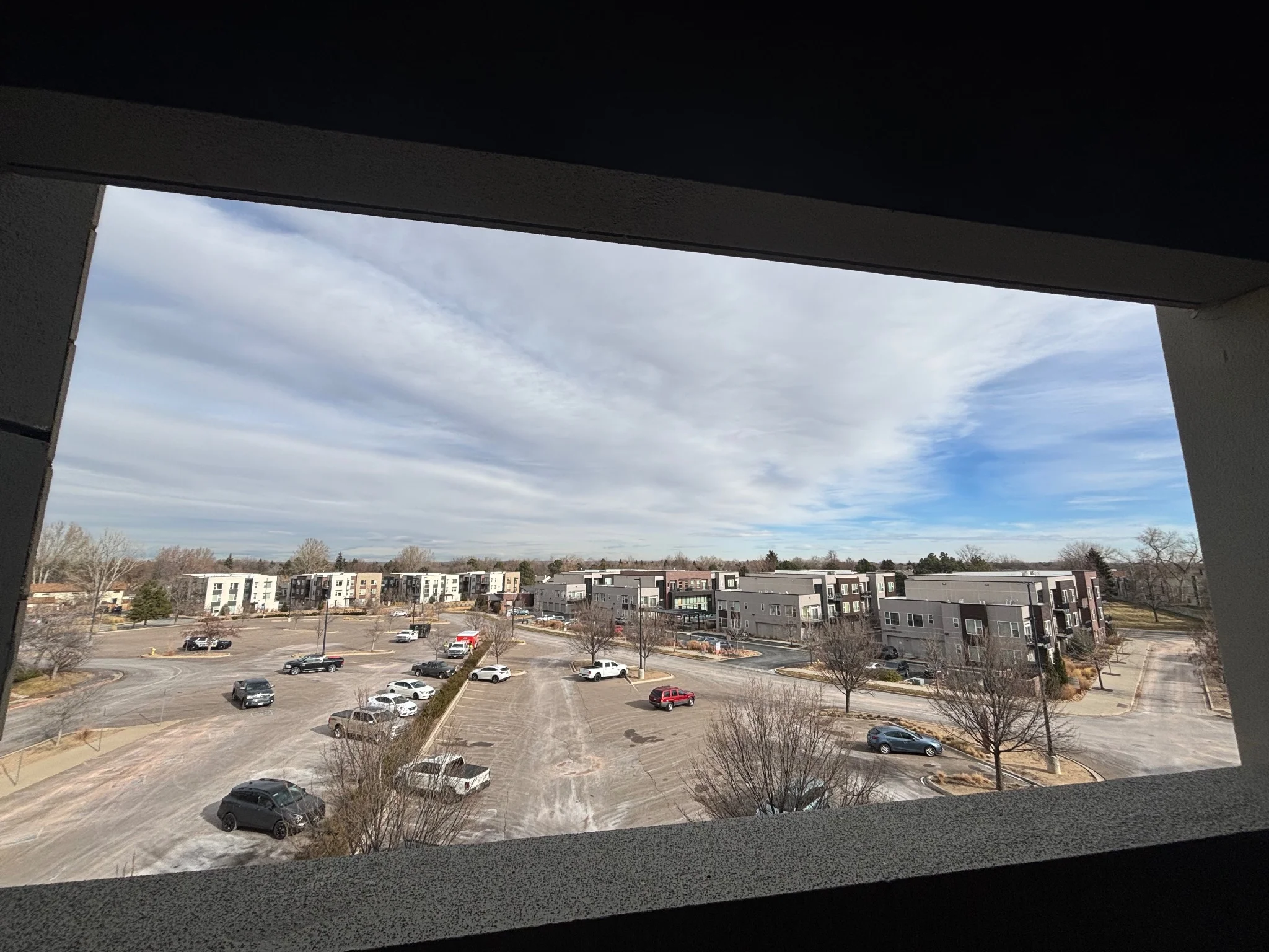 a view of a parking lot, peering out from a parking garage window, the plains beyond are quite flat and the sky is cloudy