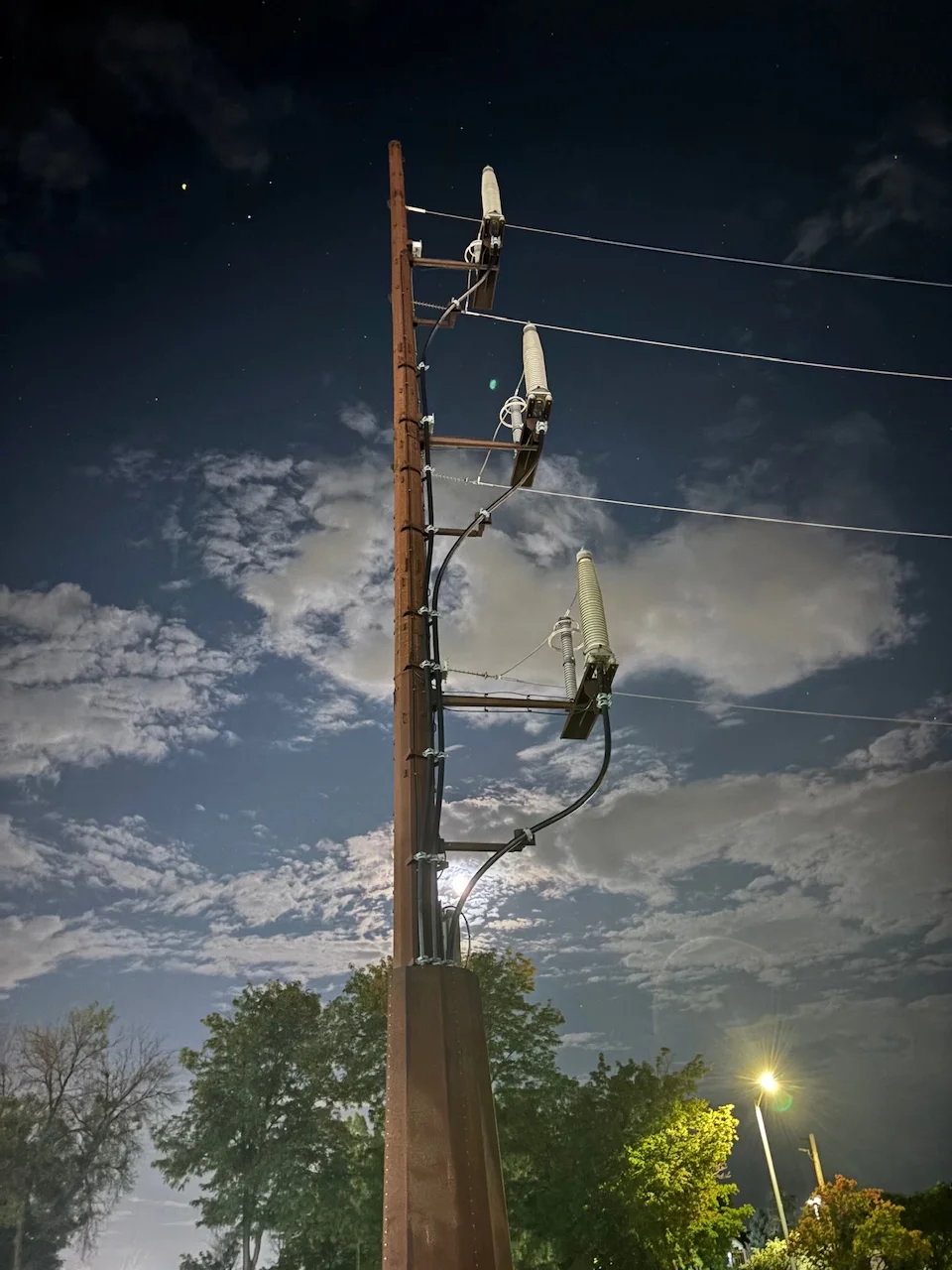a power pole, with a partly cloudy night sky in the background with the moon in between some of the pole supports
trees and a light pole can be found towards the bottom of the frame