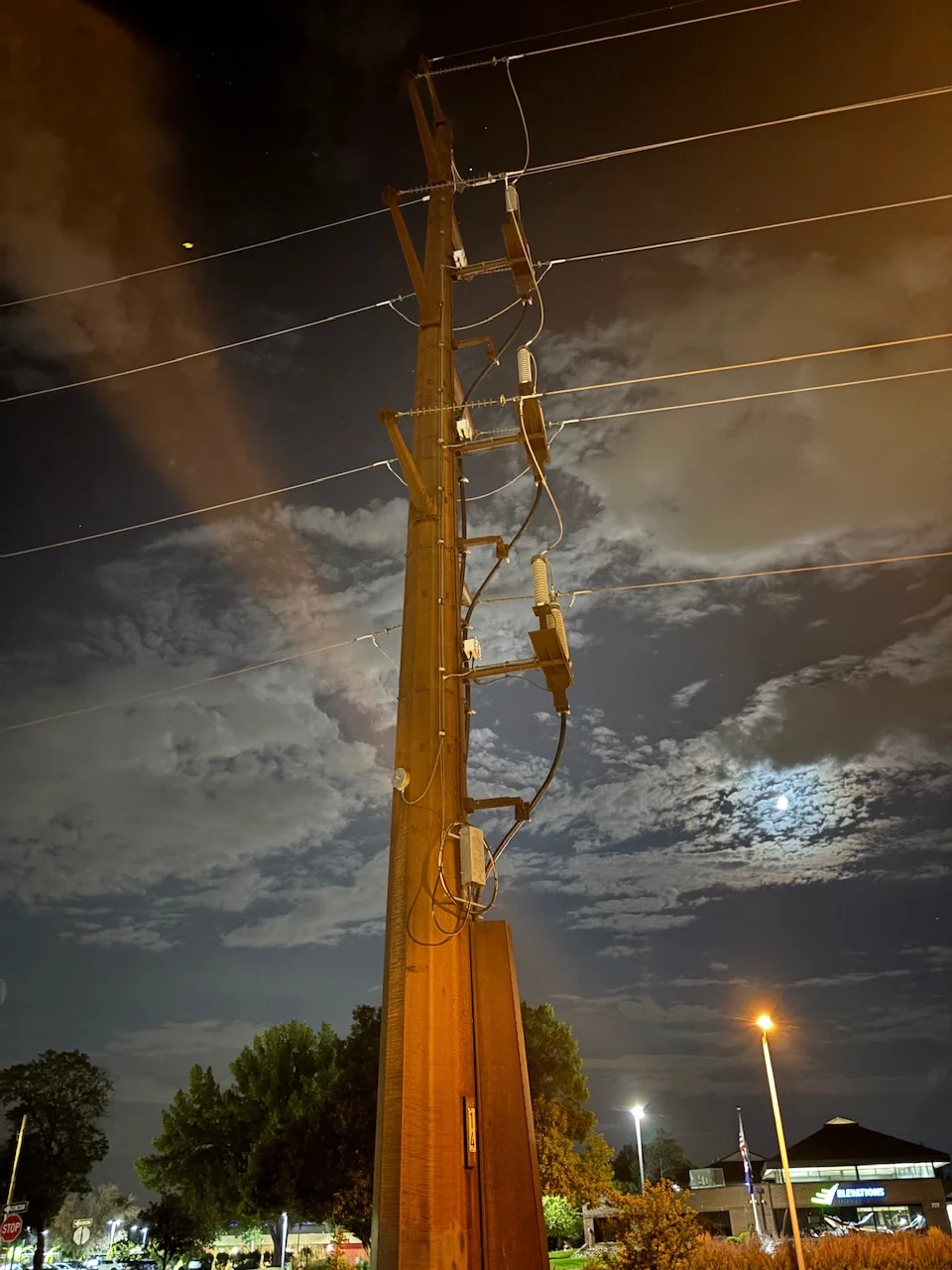 another tall power pole, this one feeling a bit more intimidating, with a partly cloudy night sky in the background with light posts and trees towards bottom frame 
of note is that there is lens flares across the frame 