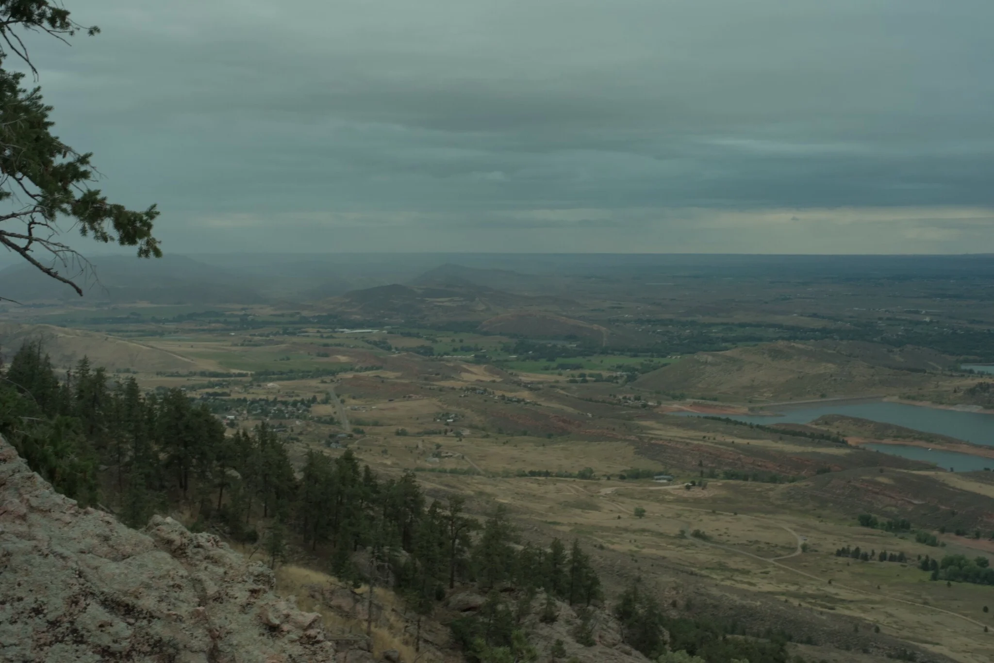 looking out onto the valley, there are pine trees nearby, and yellow fields further out... there is rain on the horizon