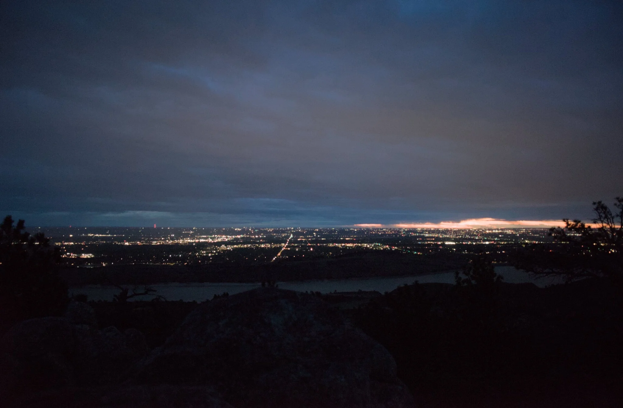 overlooking the city lights, with the sun peeking in through the clouds, silhouettes of pine trees are visible around the frame, it is morning, the air is cool and the world is waking up